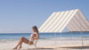 green striped beach sun shelter with girl sitting on yellow chair leaning back in pink bikini sunny sky no clouds blue water byron bay beach in background